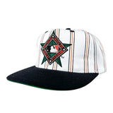 A baseball cap featuring a white front panel with thin vertical black and orange stripes, a green and black diamond-shaped 'Houston Astros' patch with MLB logo, and a black flat brim with a green underside, photographed on a light gray background.