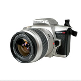 A Minolta Maxxum GT film camera with a silver body, black grip, and black lens, photographed on a light gray textured background.