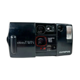 An Olympus InfinityTwin 135mm film camera in black plastic, photographed from the front against a light gray background.