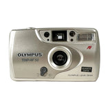 An Olympus Trip AF 50 compact camera in silver, photographed from the front on a light gray textured surface against a white background.