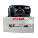 A black Pentax IQZoom 90 WR compact camera resting on top of its original gray packaging box, photographed against a neutral light gray background.