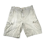 A pair of light beige cargo shorts with side flap pockets, laid flat on a white fabric background.