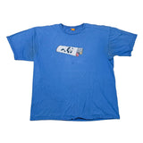 A blue short-sleeve t-shirt laid flat on a white surface, featuring a small rectangular graphic on the chest with a panda and Japanese characters. A pair of black shorts is partially visible at the bottom edge.