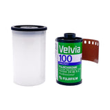 A roll of Fujifilm Velvia 100 film shown next to a clear plastic case on a white textured background.