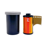 A vintage Kodachrome 64 film canister with a gray lid sits next to an open roll of film showing the gold-toned negative strip, on a white textured background.