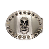 A silver-toned oval belt buckle featuring a central skull motif with blackened eye sockets and teeth, surrounded by a border of small raised studs on a white textured background.